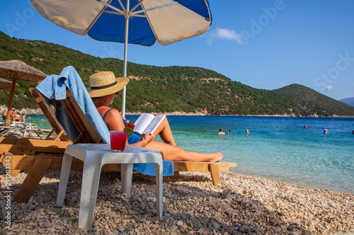 Young woman lying on a sunbed under an umbrella and reading a book next to her a table with strawberry-daiquiri cocktail on the Antisamos beach, Sami Kefalonia island, Greece.