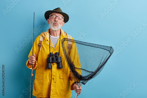 funny old man with binocular, rod and net in yellow raincaot and grren hat blowing kiss, whistling close up photo. isolated blue background, studio shot,
