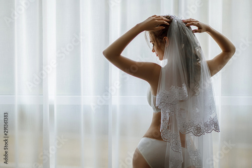 Morning bride. A beautiful girl with a white veil on her head stands near the window. Wedding portrait for the bride. Boudoir shooting. The bride in white beautiful underwear.