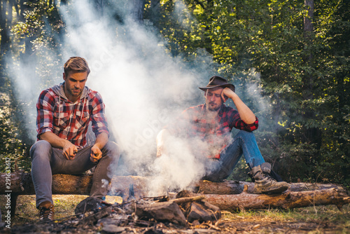 Group of friends enjoying picnic in the forest and drinking beer. Group of two male friends camping with marshmallows over a camp fire. Man doing mans job. Two friends guy camping in forest.