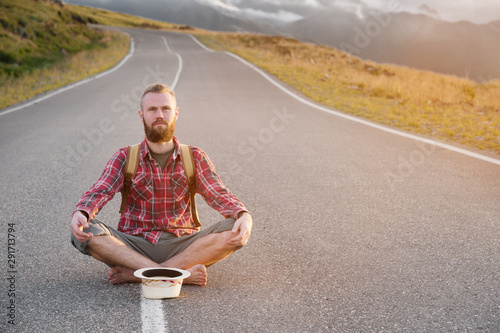Stylish barefoot bearded male hitchhiker traveler in a hat and with a backpack sits on a suburban asphalt road in the mountains at sunset and waits for cash assistance looking to the sky.