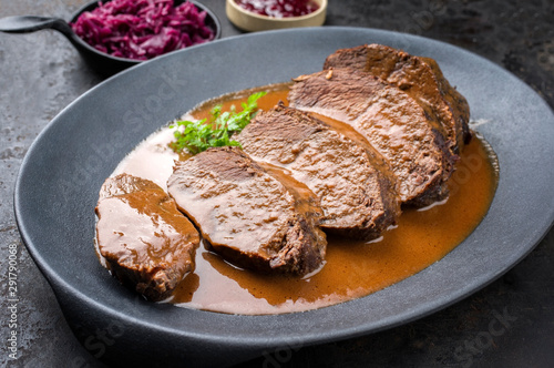 Traditional braised marinated German Sauerbraten from beef with blue kraut in spicy brown sauce as closeup in a modern design cast iron plate