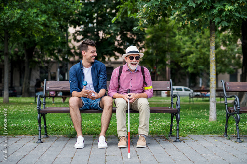 Young man and blind senior with white cane sitting on bench in park in city.