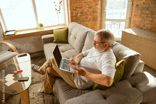 Senior man working with laptop at home - concept of home studying. Caucasian male model sitting on sofa and making notes while serfing in internet, watching cinema or webinar, online lessons.