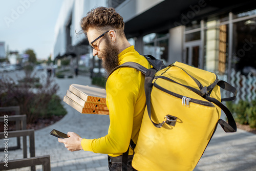 Young male courier in yellow sweatshirt delivering pizza, standing with thermal backpack and smart phone on the steret outdoors