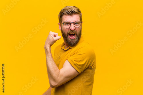 young blonde man feeling happy, satisfied and powerful, flexing fit and muscular biceps, looking strong after the gym against orange wall