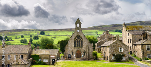 Panorama of green pastureland for sheep with drystone walls behind Congregational Church and stone cottages in Reeth North Yorkshire England