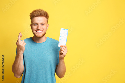 Portrait of hopeful young man with crossed fingers holding lottery ticket on yellow background, space for text