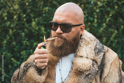 Portrait of posh chic virile bearded brutal man smoking marijuana joint, wearing brown fur gypsy style - hip hop pimp stylish guy lighting up weed (cannabis) blunt at the green background outdoors