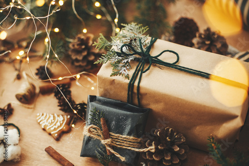 Stylish rustic christmas gifts with cedar branches and cinnamon decoration on background of golden lights, pine cones, gingerbread cookies and cotton. Merry Christmas. Wrapping gifts