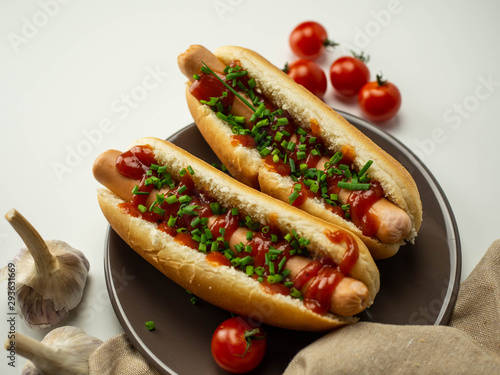 Hot dogi leżące na talerzu. Parówka, bułka, szczypiorek, pomidor. Jasne tło, widok z góry.