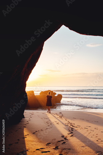 Enjoying warm summer evening sun at the beach framed by a rock cave with a female person. Sunset light. Praia da Bordeira at the Algarve Coast in Portugal, Atlantic Ocean