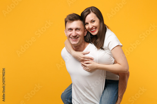 Smiling young couple two friends guy girl in white t-shirts posing isolated on yellow orange background. People lifestyle concept. Mock up copy space. Giving piggyback ride to joyful, sitting on back.