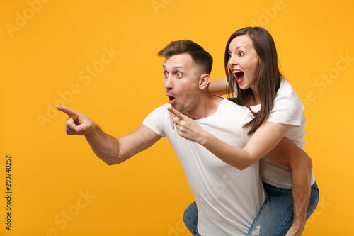 Shocked young couple in white t-shirts posing isolated on yellow orange background. People lifestyle concept. Mock up copy space. Giving piggyback ride to joyful sitting on back pointing finger aside.
