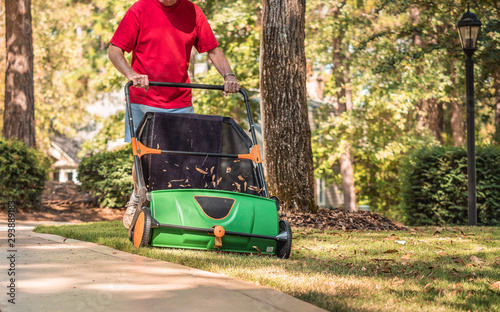 Man using manual push lawn sweeper to remove fall leaves from residential backyard grass lawn.