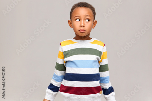 Horizontal shot of confused funny Afro American boy in striped sweater looking away with clueless facial expression, feeling guilty because he ate all sweets, pretending to be innocent. I don't know