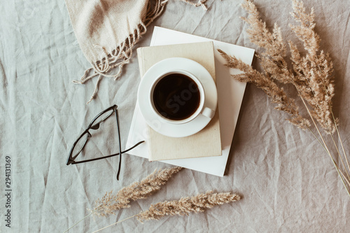 Autumn, fall composition. A cup of coffee lying on the grey linen bed with beige warm blanket, books, glasses and reeds. Lifestyle, still life concept. Flat lay, top view.