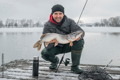 Winter fishing. Happy fisherman with pike fish at wooden platform.