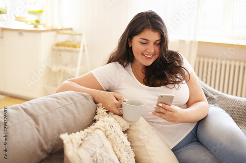 Technology, communication and leisure concept. Pretty girl with chubby cheeks relaxing at home with electronic gadget. Overweight young brunette woman texting sms on smart phone and having coffee