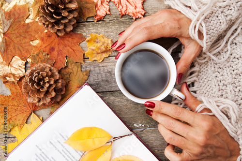 Hot coffee in the hands of a girl, a book with poems, autumn leaves, a knitted sweater on a wooden table background. Cozy autumn mood in October, November