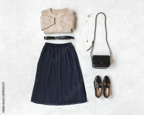 Blue midi pleated skirt, beige knitted sweater, small black cross body bag, belt, loafers (flat shoes) on grey background. Overhead view of women's casual day outfit. Flat lay, top view. Women clothes