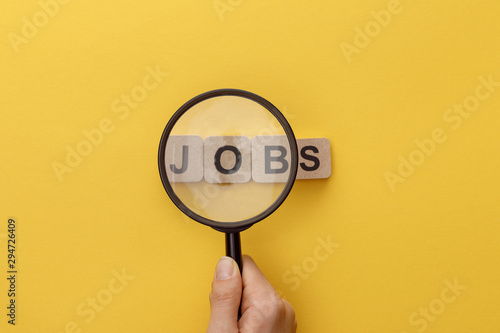 cropped view of woman holding magnifying glass under cardboard squares with jobs lettering on yellow background