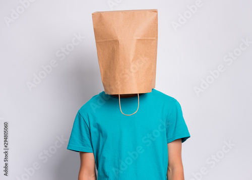 Portrait of teen boy with paper bag over head. Teenager cover head with bag posing in studio. Child pulling bag over head on gray background.