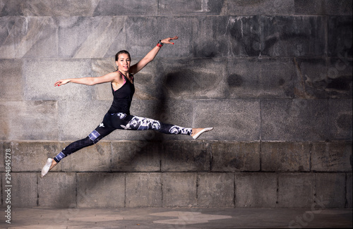 Female dancer in black tank top and leggings jumping in front of a dark gray stone wall