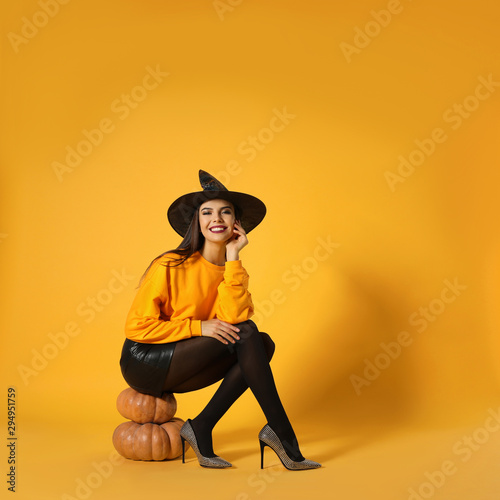 Beautiful woman wearing witch costume for Halloween party sitting on pumpkins against yellow background, space for text