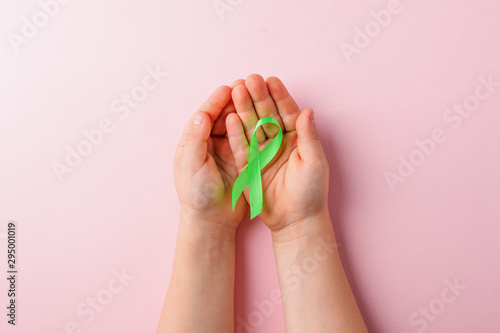 Green satin ribbon holding in kids hands on pink background. Liver, kidney cancer awareness, Glaucoma Awareness