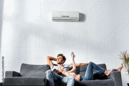 attractive girlfriend switching on air conditioner and lying on legs of handsome boyfriend
