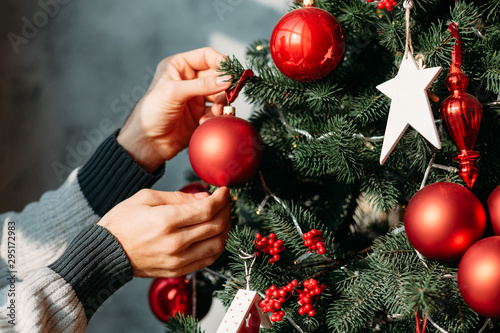 Winter holidays concept. Closeup of man hands decorating green fir tree with red ball ornaments.