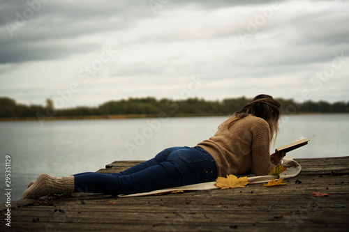 Młoda, długowosa kobieta, ubrana w ciepły sweter, jeansy i grube wełniane skarpety leży na drewnianym pomoście nad jeziorem w pochmurny jesienny dzień i czyta książkę, copyspace 