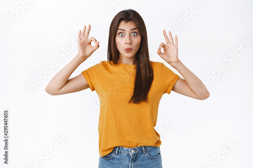 Amused, enthusiastic good-looking woman in yellow t-shirt, folding lips and pouting silly as showing her amazement and cheer, rating awesome event, show okay ok or approval sign, white background