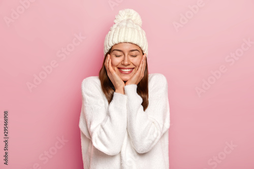 Tender relaxed carefree woman with toothy smile, wears white casual jumper and hat, feels joyful and unbothered, keeps eyes closed, poses against pink background, delighted to have devoted friends