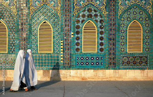 The Blue Mosque in Mazar-i-Sharif, Balkh Province in Afghanistan. Two women wearing white burqas (burkas) walk past a wall of the mosque adorned with colorful tiles and mosaics. Northern Afghanistan.