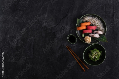 Sushi sashimi nigiri set on dark stone slate background. Seaweed wakame salad, salmon, tuna, mackerel served with sprouts, bamboo leaf, soy sauce and chopsticks. Traditional japanese food. Copy space