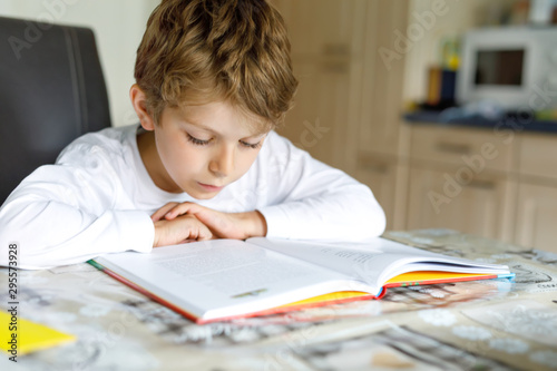 Little blonde school kid boy reading a book at home