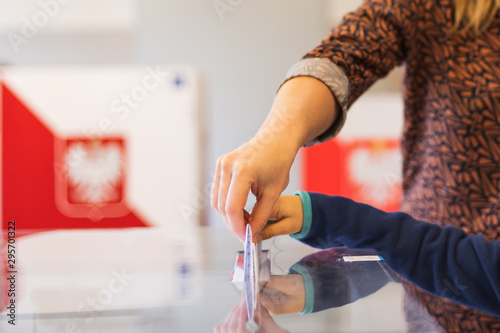 Woman's and kid's hands puting a card with a vote.to the ballot box during parliamentary elections. In the backround polish arms and flag