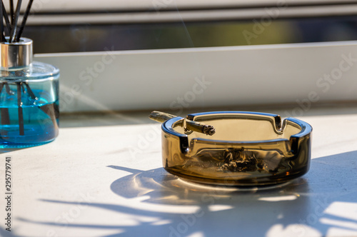 A marijuana blunt joint is burning in a glass ashtray near a window at home.