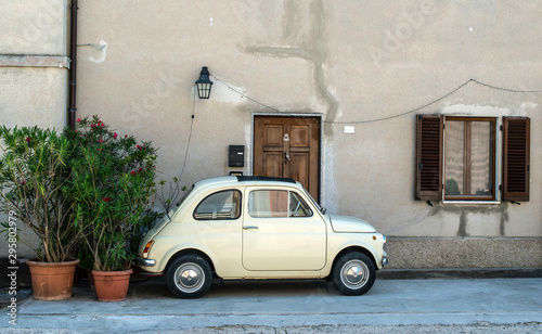 Small vintage italian car. Beige color old car in front of old house facade and flowers..