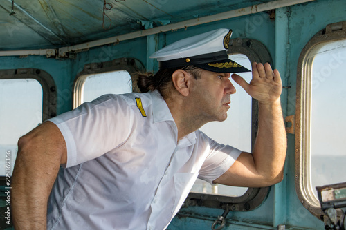 Captain standing in the wheelhouse and looking to ahead. A sailor in the white uniform looking to sea from control room - navigation bridge.