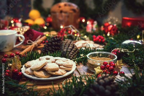 Christmas ingredients table with a cookies in the center on a wooden board, accompanied by orange, panettone, cinnamon, pine. Festive pine decoration on red rustic background