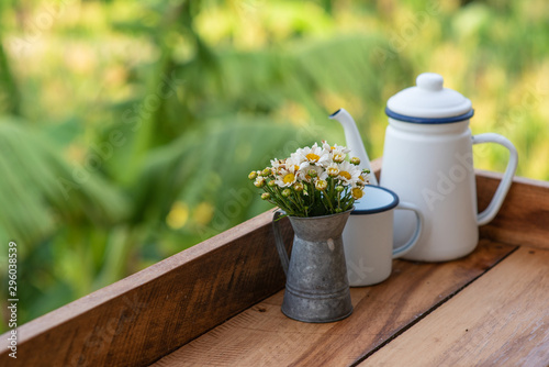 The flower vase has white-yellow flowers placed beside the teapot on the wooden floor.
