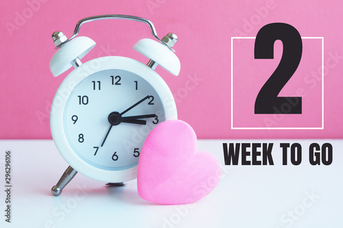 A small white alarm clock and a pink heart figurine. The inscription "2 week to go"...