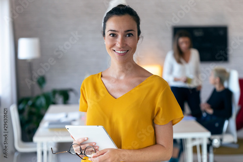 Pretty young businesswoman looking at camera. In the background, her colleagues working in the office.