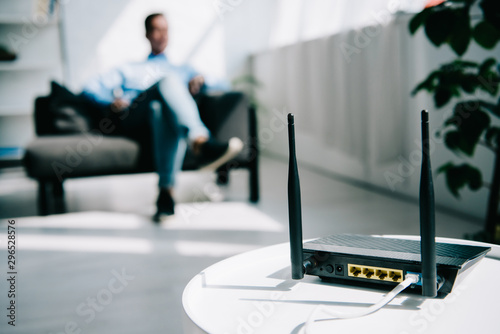 selective focus of black plugged router on white table and businessman sitting on sofa