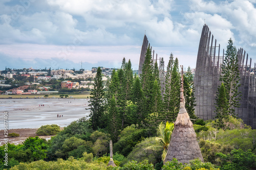 Tjibaou Cultural Centre, the Kanak native art museum with a traditional straw hut in foreground and the city of Noumea in background and people walking on sea shore at low tide in New Caledonia.