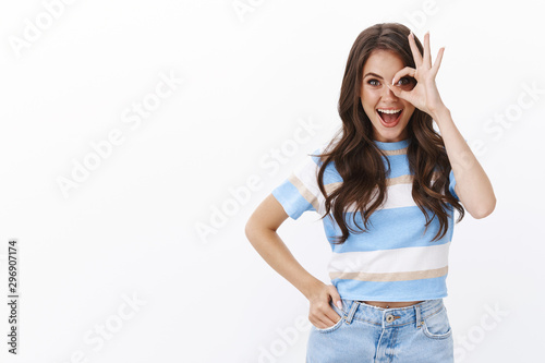 Excited cheerful glamour european woman with curly haircut wear t-shirt and jeans, show okay approval sign, smiling amused and joyful, contemplate cool event, look through fingers astonished