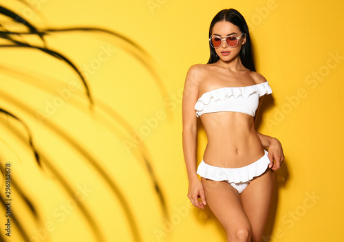 Beautiful young woman in white bikini with sunglasses on yellow background. Space for text